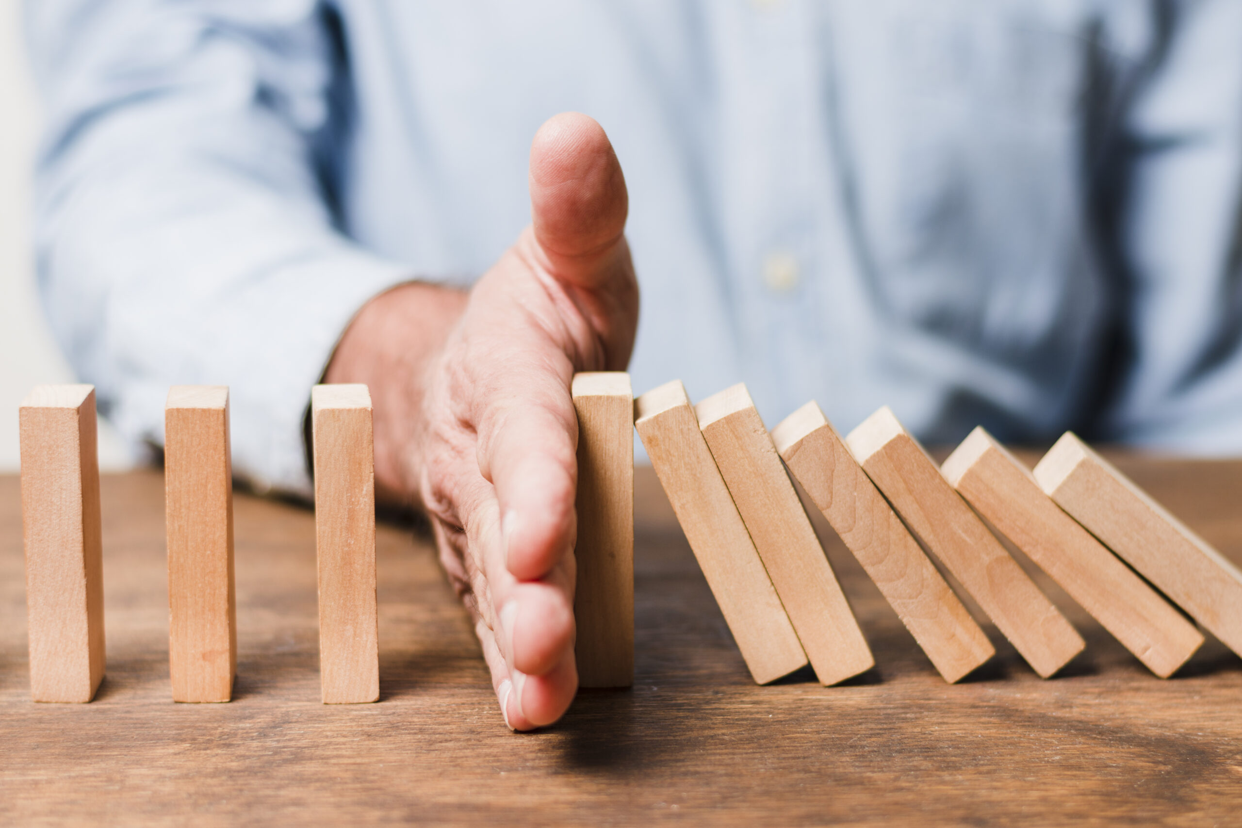 A hand in a blue shirt stops a line of falling wooden dominoes on a wooden table, symbolizing intervention and control.